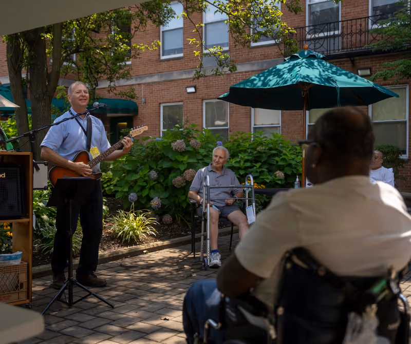 An outdoor courtyard at Chapin Home for the Aging where a man is playing an electric guitar and singing into a microphone. Several elderly people are seated nearby, including one man with a walker and another person in a wheelchair, enjoying the music. The area is shaded by trees and green umbrellas, with a brick building in the background.
