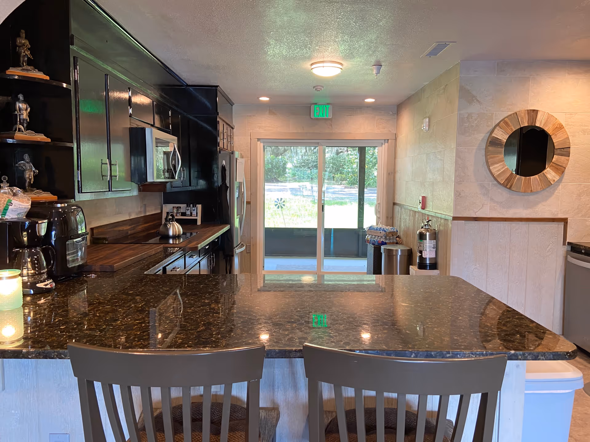 Interior view of a kitchen area with dark wooden cabinets, a granite countertop island with two chairs, a microwave, a refrigerator, and a sliding glass door leading outside. There is a round wooden-framed mirror on the wall and a fire extinguisher near the door.