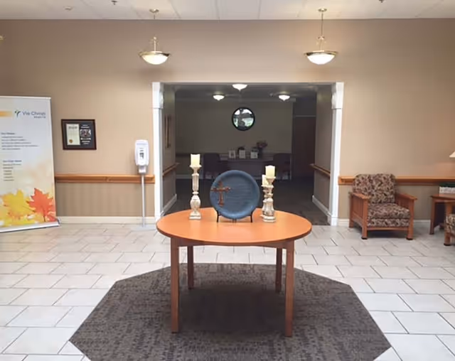 A lobby area with a round wooden table in the center displaying two candle holders and a decorative plate with a cross. The floor is tiled with a dark rug under the table. To the left, there is a standing banner with the Via Christi logo and text, and a hand sanitizer dispenser mounted on the wall. To the right, there are two upholstered armchairs and a small wooden table with a lamp. The walls are beige with a wooden chair rail, and there is an open doorway leading to a dimly lit room with a circular window.