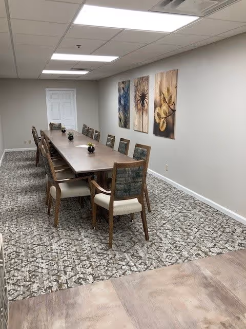 Long rectangular dining/meeting room with a wooden table surrounded by chairs, patterned carpet, and three abstract artworks on the wall.