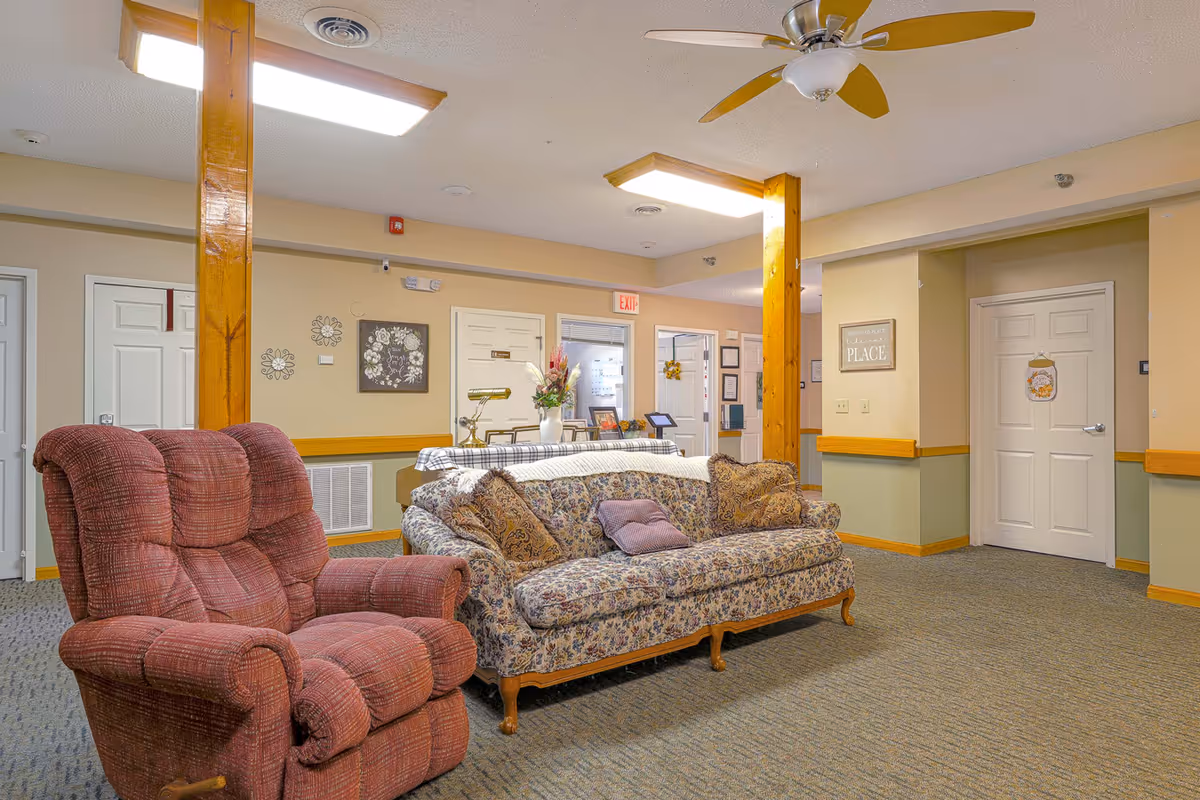 A cozy living room area in a senior living facility featuring a floral patterned sofa with multiple cushions and a red upholstered recliner chair. The room has beige walls with wooden trim and support beams, a ceiling fan with wooden blades, and fluorescent ceiling lights. There are several doors and decorative wall art, including a sign that reads 'There is no place like this PLACE.'