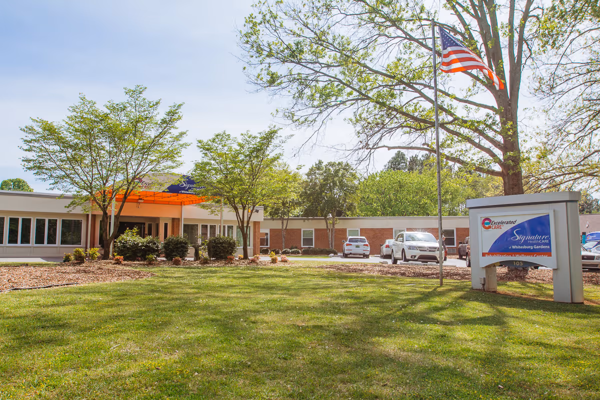 Exterior view of a single-story healthcare facility with a green lawn and trees in front. There is an American flag on a flagpole and a sign near the entrance displaying the name 'Excelerated Care Signature Healthcare Whitesburg Gardens'. Several cars are parked in the parking lot beside the building.