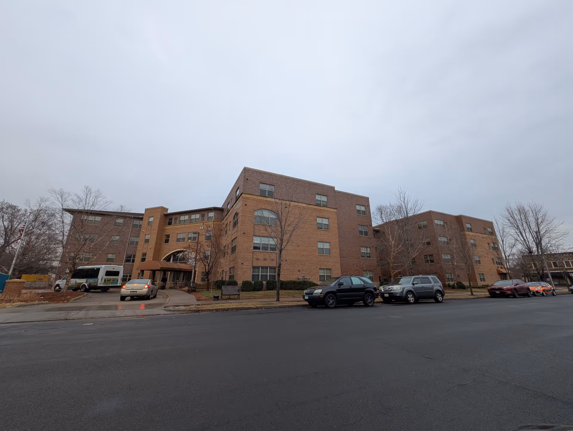 Exterior view of a multi-story brick building with several windows, a covered entrance, and a driveway with cars and a shuttle bus parked. Leafless trees and a bench are visible on the lawn in front of the building under an overcast sky.