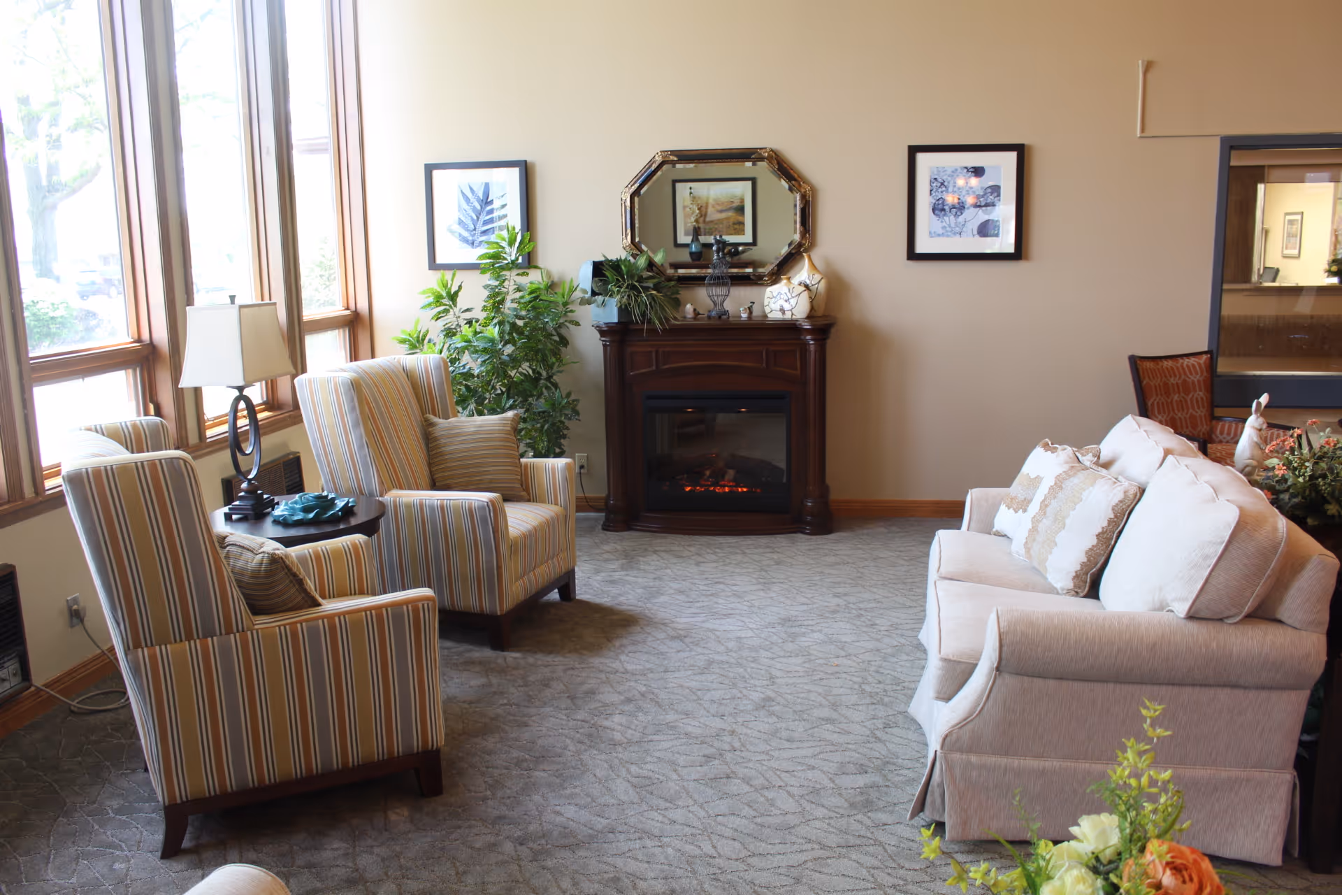 Sunlit sitting area with striped armchairs, a sofa, a fireplace console, plants, and framed artwork.