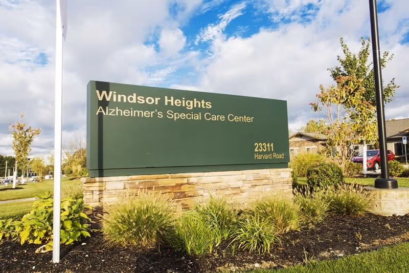 Outdoor green sign reading "Windsor Heights Alzheimer's Special Care Center" with address in front of landscaping and a partial view of the building.