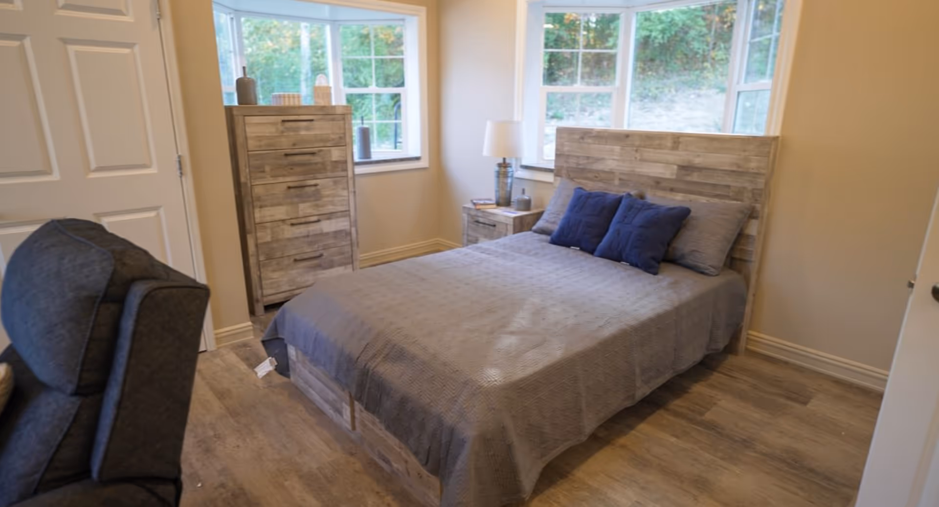 A cozy bedroom with a wooden bed frame and headboard, covered with a gray bedspread and two blue pillows. There is a matching wooden dresser and nightstand, with a lamp and decorative items on top. The room has two large windows letting in natural light and a gray upholstered chair partially visible in the foreground.