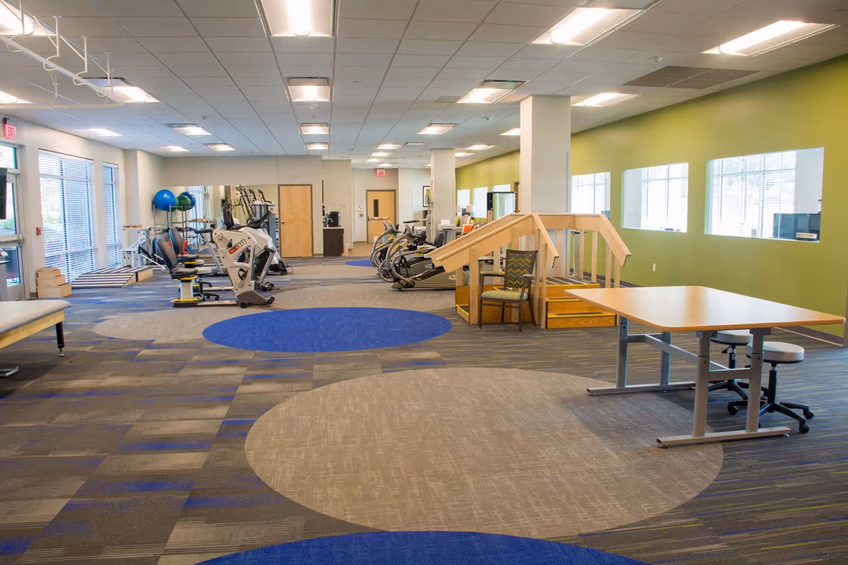 A spacious rehabilitation or fitness room with exercise equipment including stationary bikes and treadmills, a wooden ramp with handrails, chairs, and tables. The room has large windows along one wall, green painted walls, and a patterned carpet with blue and beige circular designs.