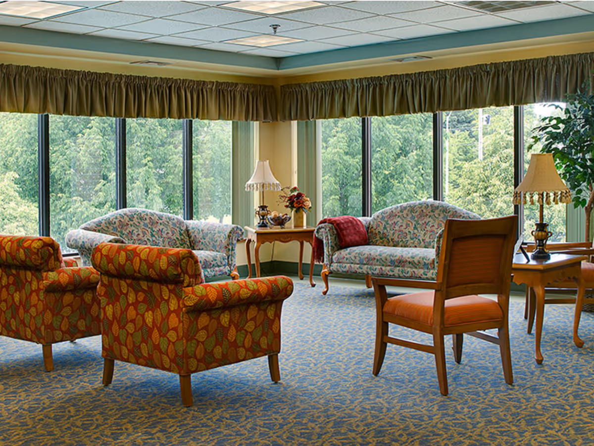 A bright and cozy living room area with large windows showing green trees outside. The room features two floral-patterned sofas, two armchairs with a leaf pattern, a wooden chair with an orange cushion, two side tables with lamps, and a small flower arrangement on one table. The carpet has a blue and gold pattern, and the windows are adorned with green valances.