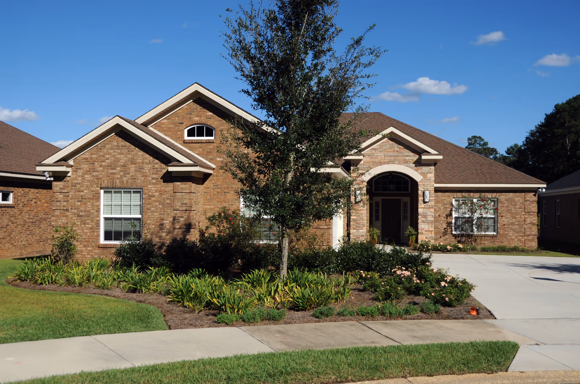 Front view of a brick single-story house with a landscaped garden, tree, and driveway under a blue sky.