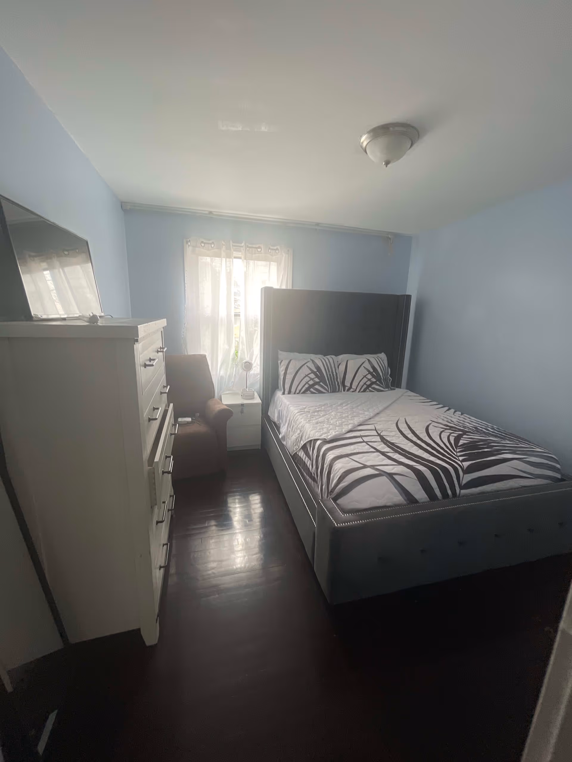 A small bedroom with light blue walls and dark wooden floor. The room contains a gray upholstered bed with patterned bedding, a white chest of drawers with a TV on top, a brown armchair, and a white nightstand with a small lamp. A window with sheer white curtains lets in natural light.