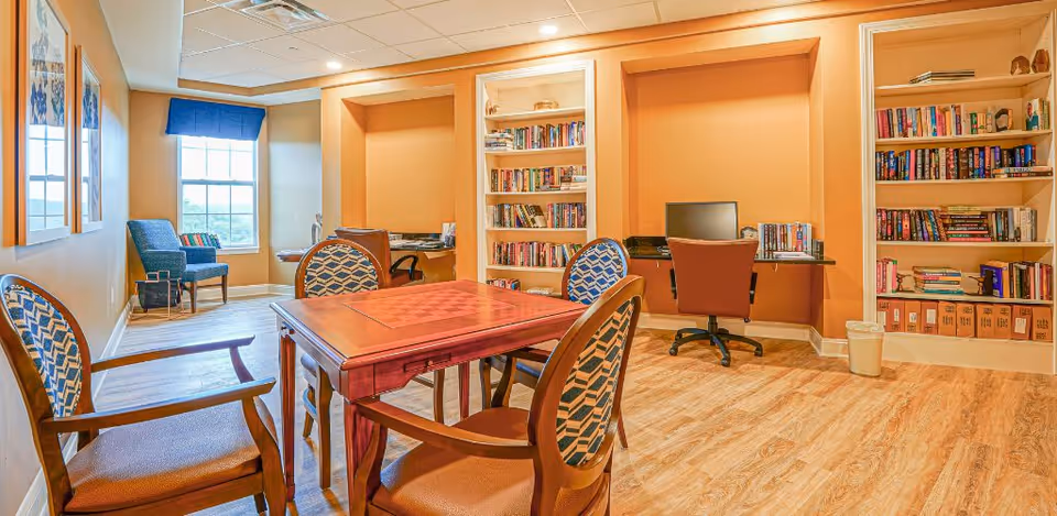 A cozy interior room with a wooden table and four chairs in the center, two computer desks with chairs and desktop computers against the far wall, and bookshelves filled with books. There is a window with a blue valance and an armchair near it. The room has warm yellow walls and wood flooring.