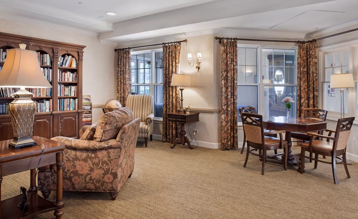 A cozy living room area in a senior living facility featuring a floral patterned sofa, a striped armchair, a wooden bookshelf filled with books, a globe, and a wooden table with four chairs near windows with floral curtains. Several lamps provide warm lighting.