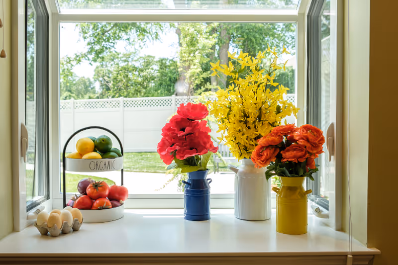 A bright windowsill with three colorful vases holding artificial flowers in red, yellow, and orange. To the left, a two-tiered stand labeled 'ORGANIC' holds lemons, limes, and tomatoes. A carton with six eggs is also placed on the windowsill. Outside the window, a white fence and green trees are visible.