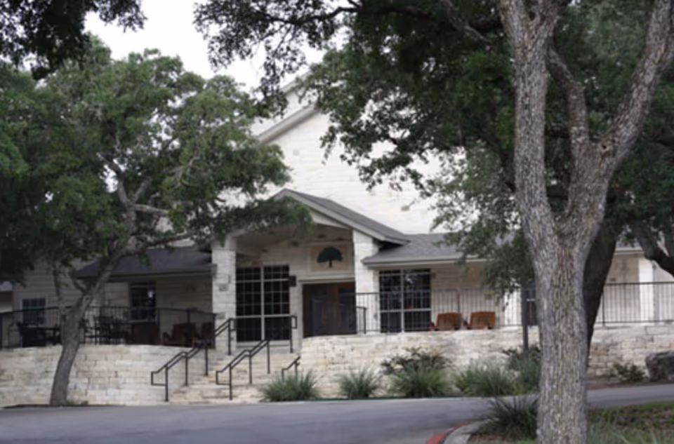 Exterior view of a stone building with a covered porch, surrounded by trees and greenery. The building has large windows and a railing along the porch with steps leading up to the entrance.