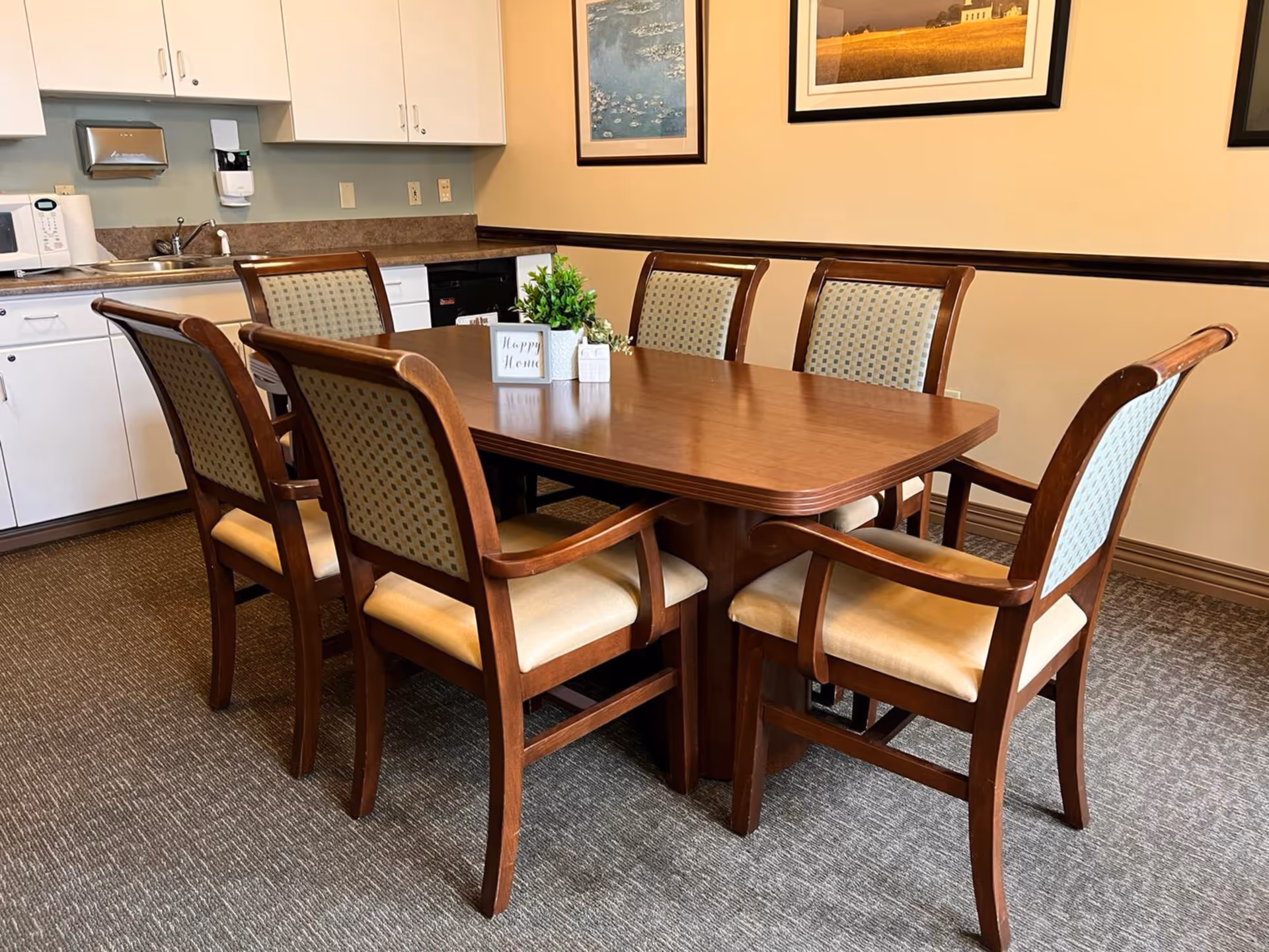 A dining area with a wooden table surrounded by six cushioned chairs with patterned backs. The room has a carpeted floor and beige walls adorned with framed artwork. In the background, there is a kitchenette with white cabinets, a microwave, a sink, and a dishwasher. On the table, there is a small plant and a decorative sign that reads 'Happy Home'.