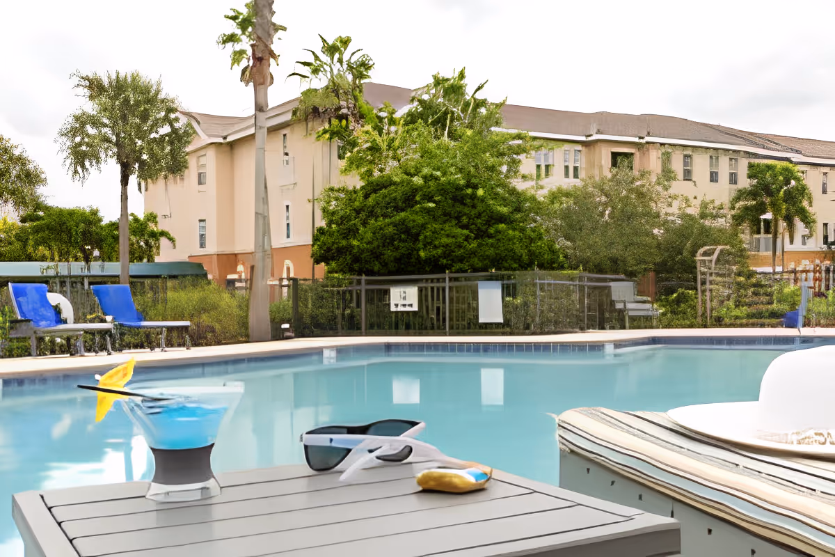 Outdoor swimming pool with lounge chairs and a table holding a blue cocktail, sunglasses, and a white hat. In the background, there are trees, bushes, and a multi-story building under a cloudy sky.