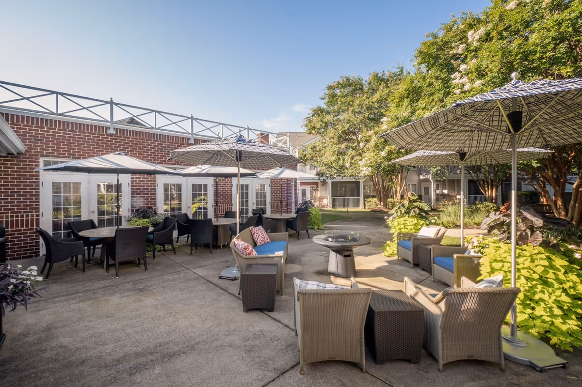 Outdoor patio area at Chancellor's Village with multiple seating arrangements including wicker chairs and tables under large patterned umbrellas. The patio is surrounded by greenery and trees, with a brick building featuring multiple French doors in the background.
