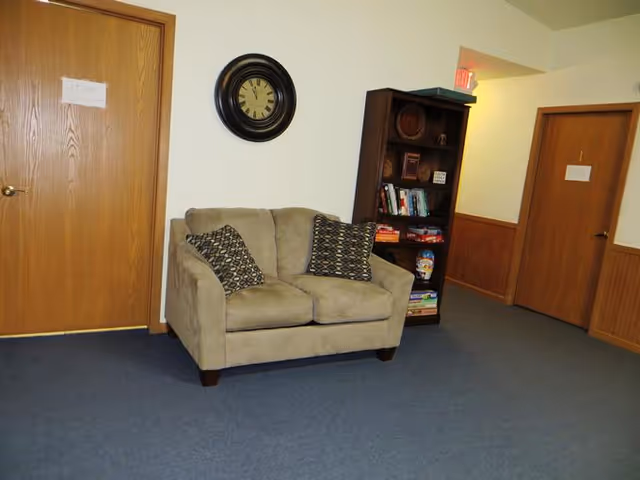 Small seating area with a beige loveseat, two patterned pillows, a wall clock, and a bookshelf between two wooden doors in a carpeted hallway.