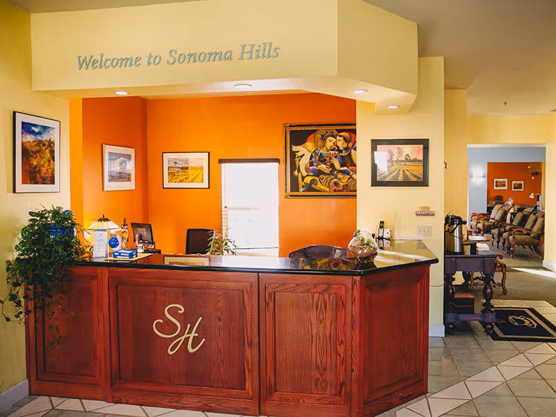 Reception desk area at Sonoma Hills Retirement Living Community with a wooden counter featuring the initials 'SH'. The wall behind the desk is painted orange and decorated with framed artwork. A plant and various office items are on the desk. The surrounding walls are light yellow, and there is a seating area visible in the background.