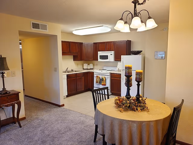 Interior view of a senior living facility showing a small dining area with a round table covered with a beige tablecloth, decorated with three candles and a floral arrangement. The dining area has two black chairs and is adjacent to a kitchen with dark wooden cabinets, a white refrigerator, microwave, stove, and dishwasher. The floor transitions from carpet in the dining area to tile in the kitchen. A small wooden side table with a lamp is visible on the left side.