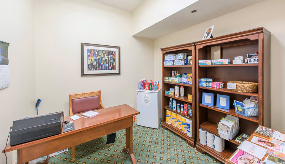 Small interior office/storage room with a desk and chair, a printer, mini fridge, and two wooden shelves stocked with supplies.