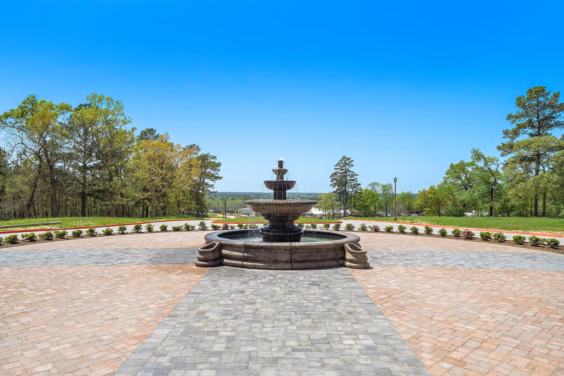 Outdoor view of a multi-tiered stone fountain centered on a paved circular area with a pathway leading up to it, surrounded by green grass, small bushes, and tall trees under a clear blue sky.