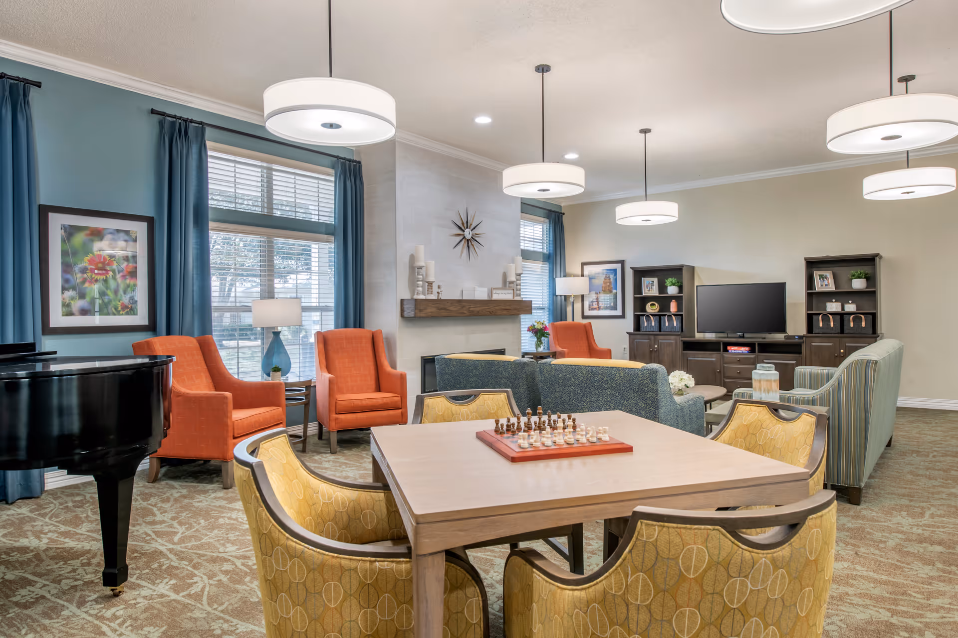 A cozy senior living facility common area with a square table and four yellow patterned chairs in the foreground, a chessboard on the table, a black grand piano to the left, two orange armchairs near large windows with blue curtains, a fireplace with decorative candles and a clock above it, and a TV with dark wood cabinets and shelves in the background. The room is well-lit with modern circular pendant lights and has a carpeted floor.
