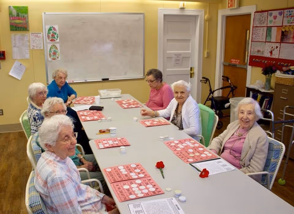 A group of elderly women sitting around a long table playing bingo in a well-lit room with yellow walls. The table has bingo cards and white bingo chips. There are walkers and a bulletin board with colorful papers on the wall in the background.