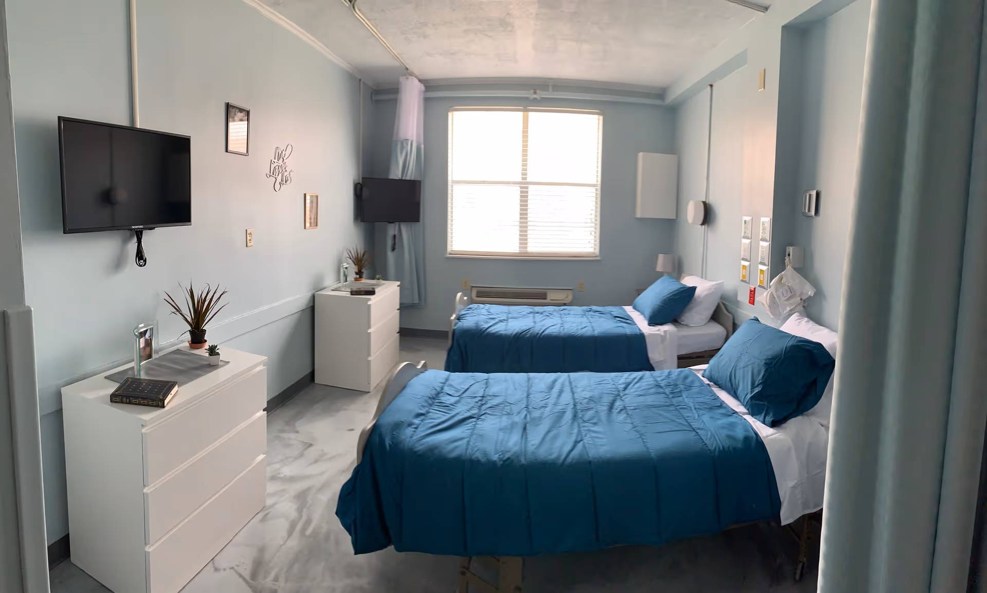 A nursing facility room with two single beds made with blue bedding and white pillows. Each bed has a small bedside lamp and medical equipment mounted on the wall behind it. There are two white dressers, each with a small plant and a book on top. Two flat-screen TVs are mounted on the walls. A large window with blinds lets in natural light, and the walls are painted light blue.