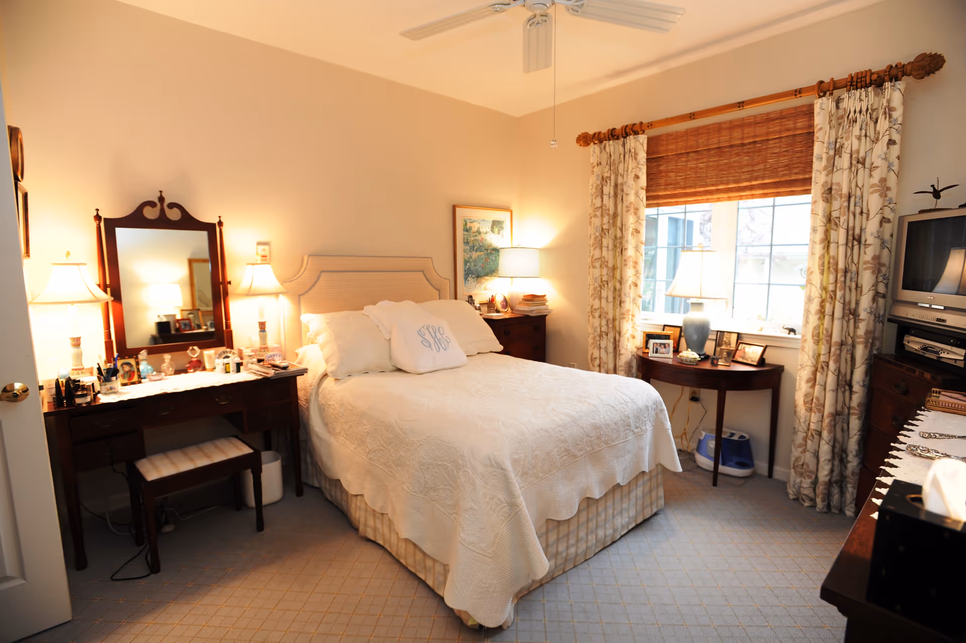 A cozy bedroom with a neatly made bed covered in a white quilt and pillows. On the left side of the bed is a wooden vanity table with a mirror, two lamps, and various personal items. To the right of the bed is a wooden nightstand with a lamp and books. A window with floral curtains and a bamboo shade lets in natural light. A small round table beneath the window holds framed photos and a lamp. A TV and other furniture are visible on the right side of the room. The room has a ceiling fan and light-colored carpet.