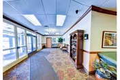 Carpeted lobby hallway with seating, display shelves, potted plants and large windows at a senior living facility.