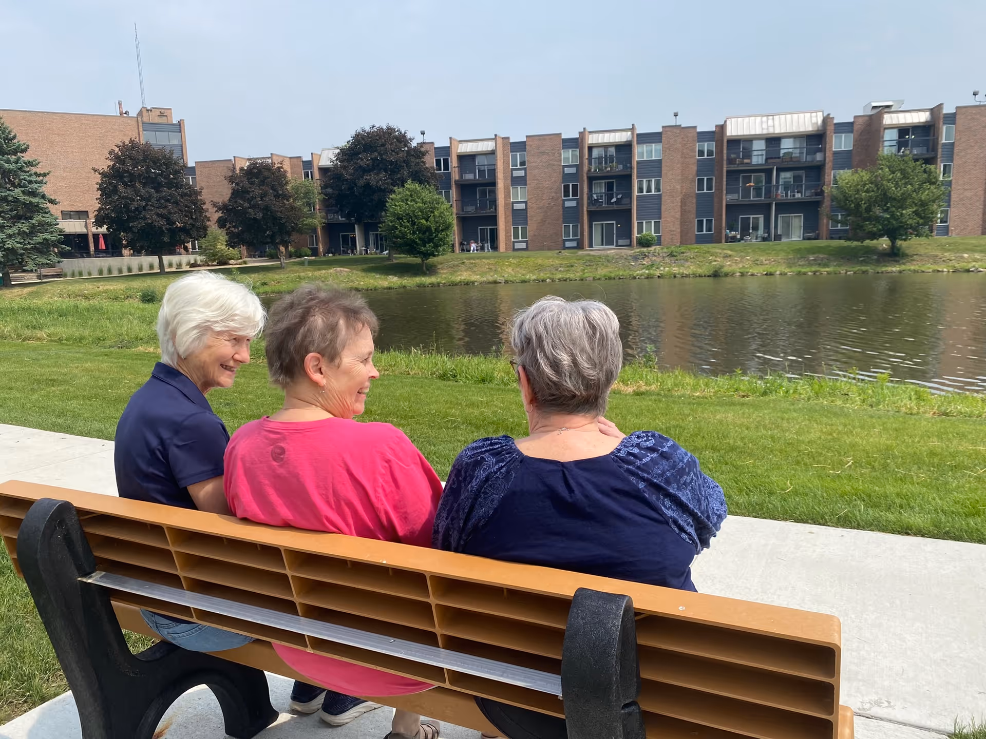 Three elderly women sitting on a wooden bench by a pond, with a multi-story brick building and trees in the background under a clear sky.