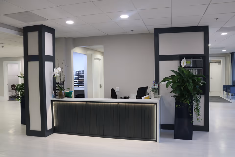 Reception desk area inside a senior living facility with a modern design. The desk has a dark front panel with subtle lighting underneath the countertop. Behind the desk is a computer monitor and office chair. The area is decorated with potted plants and flowers. The walls and floor are light-colored, and there are doorways and seating visible in the background.