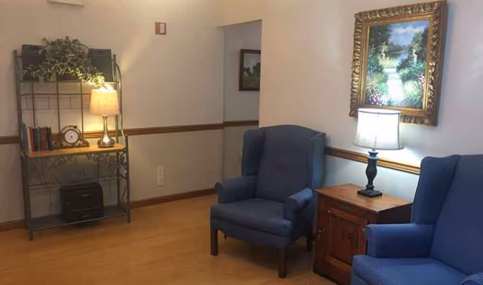 A small sitting area with two blue armchairs, a wooden side table and lamp, framed artwork, and a shelving unit with a lamp and decor.