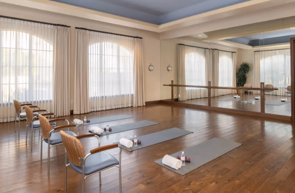 A spacious exercise or yoga room with wooden floors, large windows with sheer curtains, and a wall-length mirror. Four exercise mats are laid out on the floor, each with a rolled towel, a small water bottle, and a pair of light dumbbells. Four chairs with wooden armrests and blue cushions are positioned next to the mats. The room is well-lit with natural light and has a calm, inviting atmosphere.
