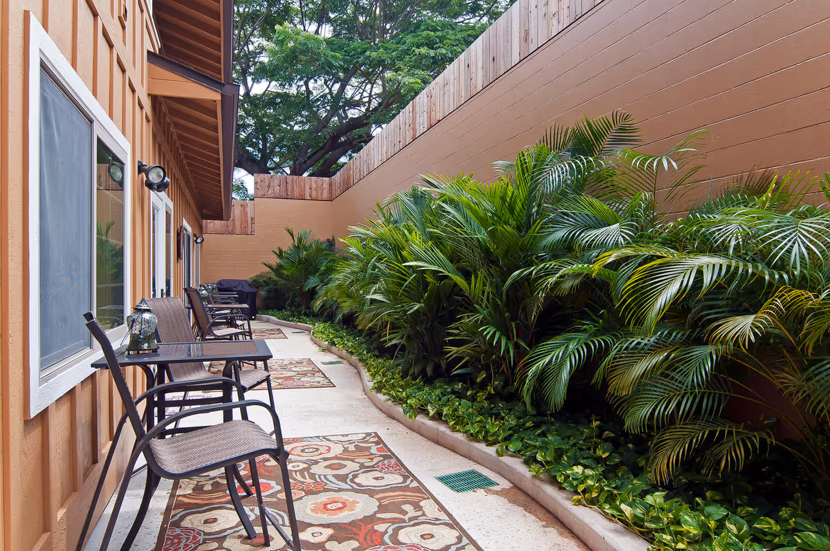 Outdoor patio area with a row of metal chairs and small tables along the side of a building. The ground is decorated with patterned rugs, and there is a garden bed filled with lush green plants and palm-like foliage next to a tall beige wall. Trees are visible above the wall in the background.