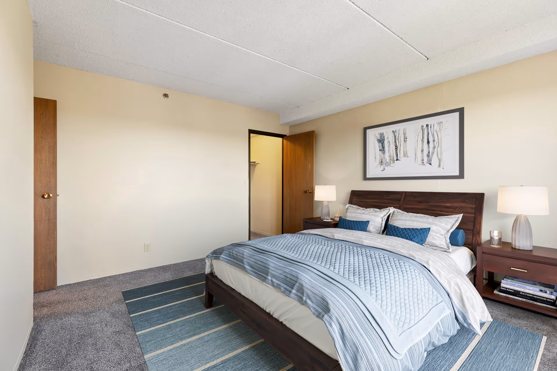 Well-lit bedroom with a wooden bed dressed in blue bedding, two nightstands with lamps, framed art above the headboard, and an open closet door.