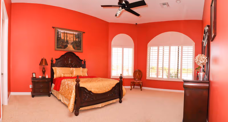 A bedroom with red walls and beige carpet featuring a dark wooden bed with gold bedding and red blanket. There is a matching nightstand with a lamp and picture frame, a wooden chair near two large windows with white shutters, and a dark wooden dresser with a mirror and decorative items. A ceiling fan is mounted on the white ceiling.