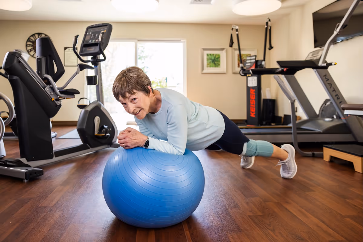 An elderly woman in athletic wear is exercising on a blue stability ball in a fitness room with various exercise machines including a treadmill and stationary bike. The room has wooden floors, light-colored walls, and framed pictures on the wall.