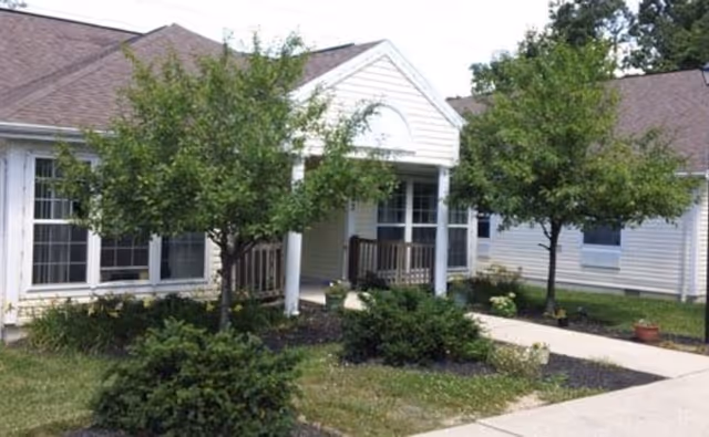 Front entrance of a single-story residential building with a covered porch, walkway, and small trees and shrubs.