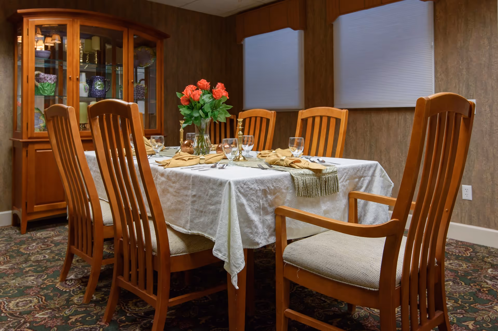 A dining room with a rectangular table covered with a white tablecloth and set with glasses, napkins, and silverware. There is a vase with red roses and two brass candlesticks as centerpieces. Six wooden chairs with beige cushions surround the table. In the background, there is a wooden china cabinet with glass doors displaying decorative items. The room has patterned carpet and two windows with closed blinds.