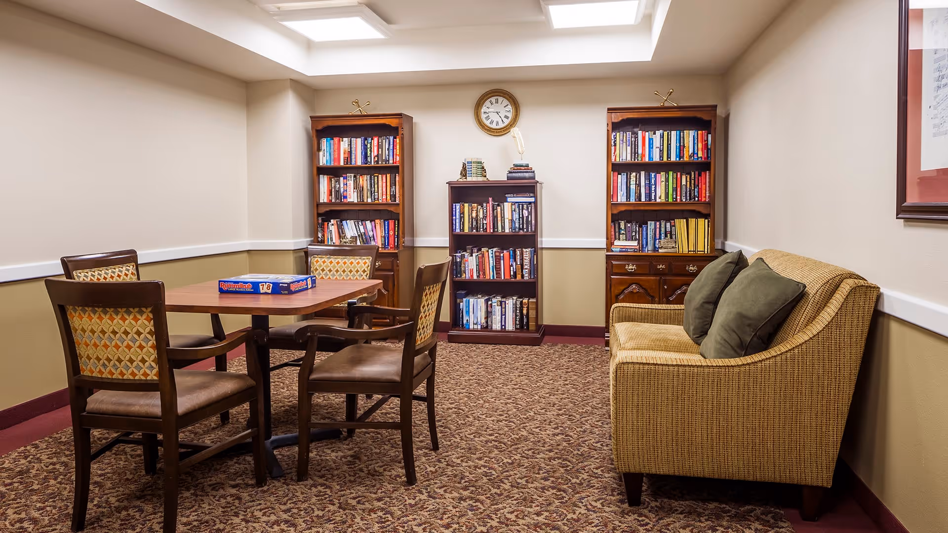 A cozy room with a square wooden table surrounded by four chairs with patterned cushions. On the table is a Rummikub game box. Behind the table are three wooden bookshelves filled with books, and a round clock is mounted on the wall above the center bookshelf. To the right is a cushioned loveseat with two dark green pillows. The room has beige walls with a white chair rail and patterned carpet flooring.