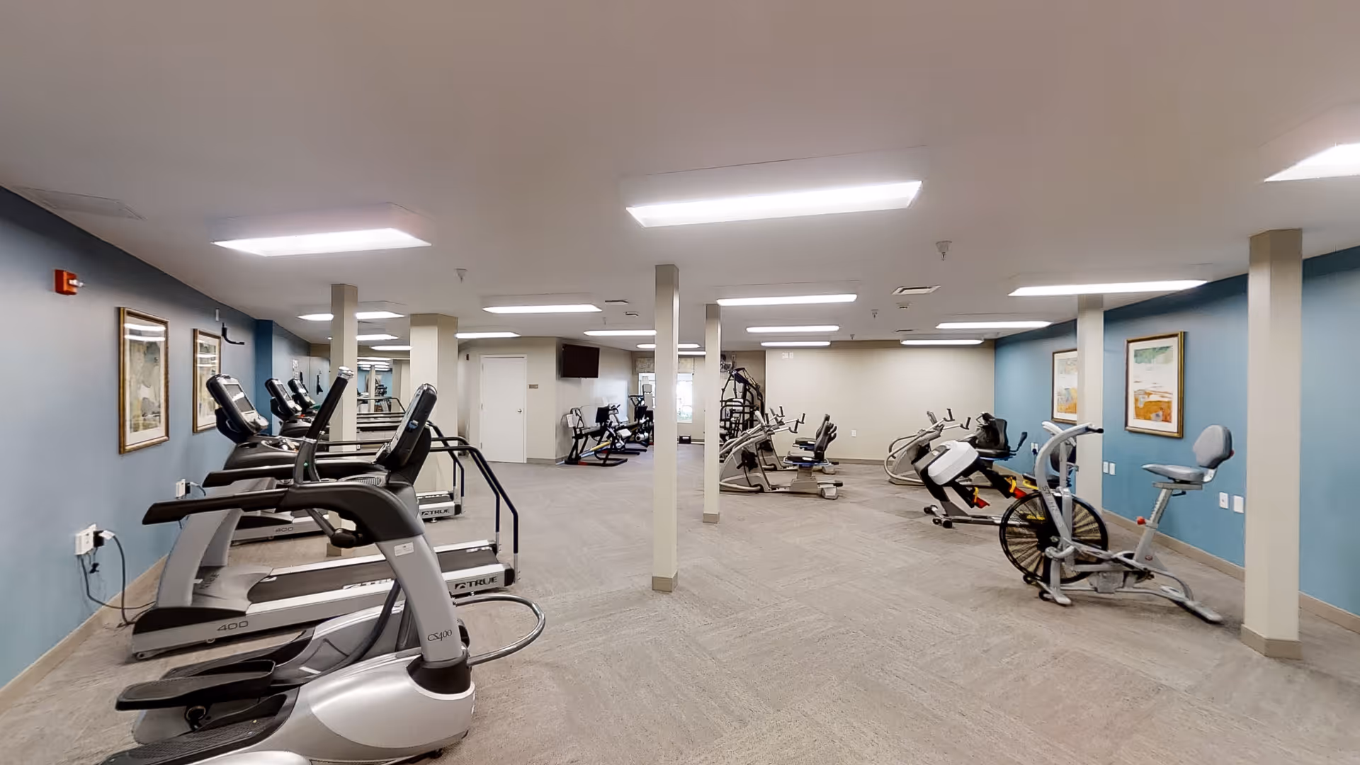A spacious fitness room with multiple exercise machines including treadmills, stationary bikes, and strength training equipment. The room has light gray carpeted floors, blue and beige walls adorned with framed artwork, and bright overhead lighting.