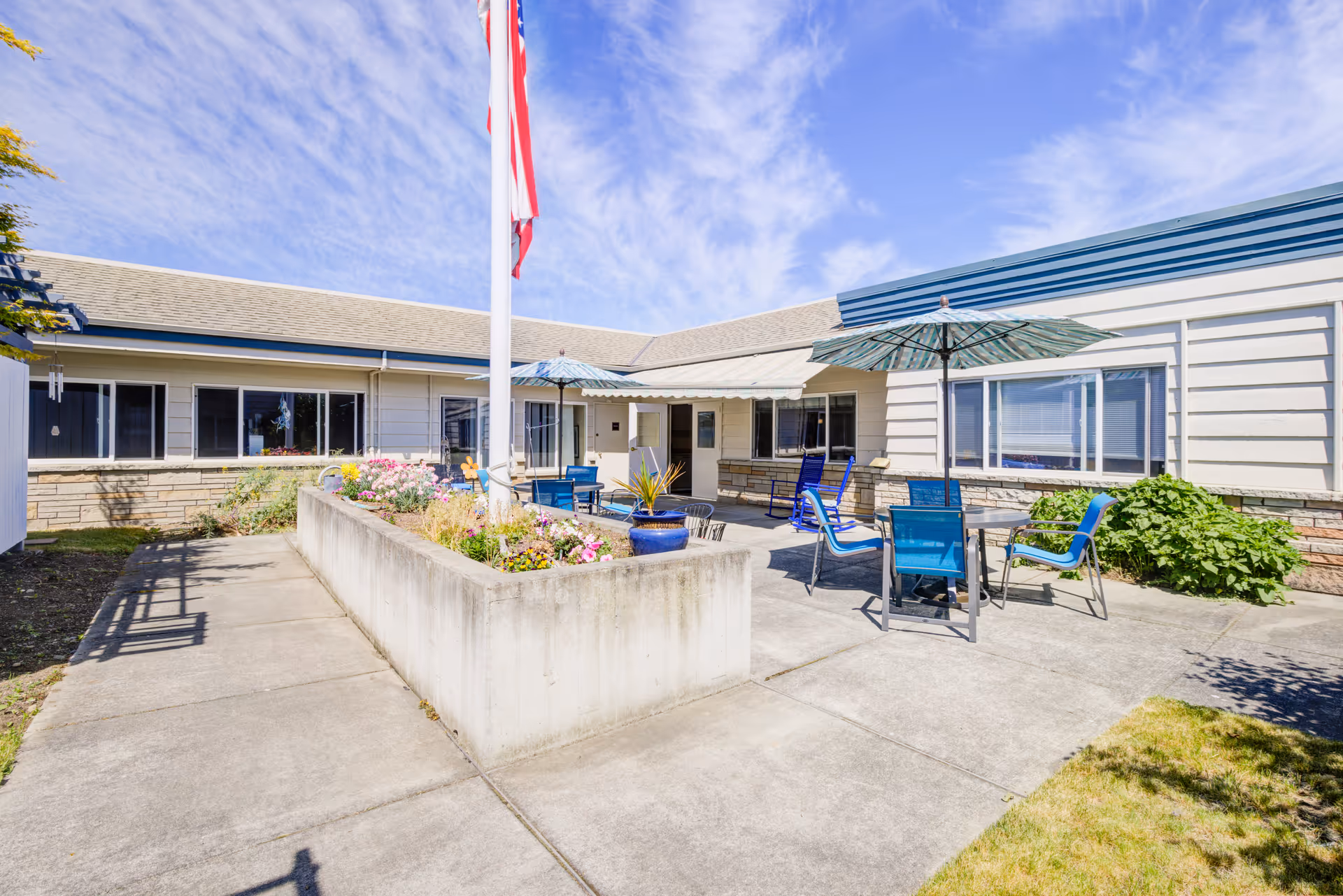 Outdoor courtyard area at The Cottages of Sequim featuring a concrete walkway, a raised flower bed with colorful flowers, several blue chairs and tables with umbrellas, and a flagpole with an American flag under a partly cloudy sky.