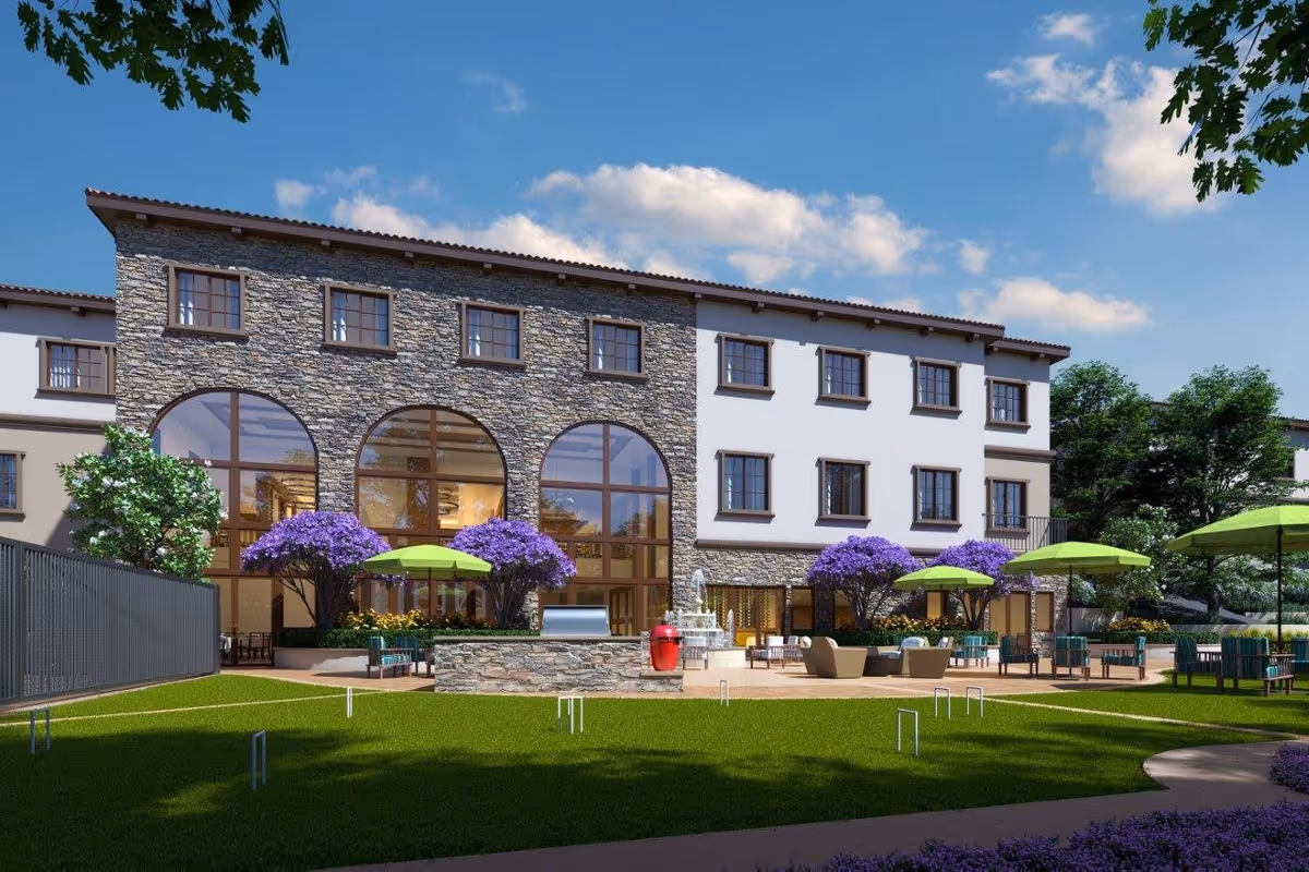 Stone-and-stucco building with large arched windows, outdoor patio seating, green umbrellas and a landscaped lawn.