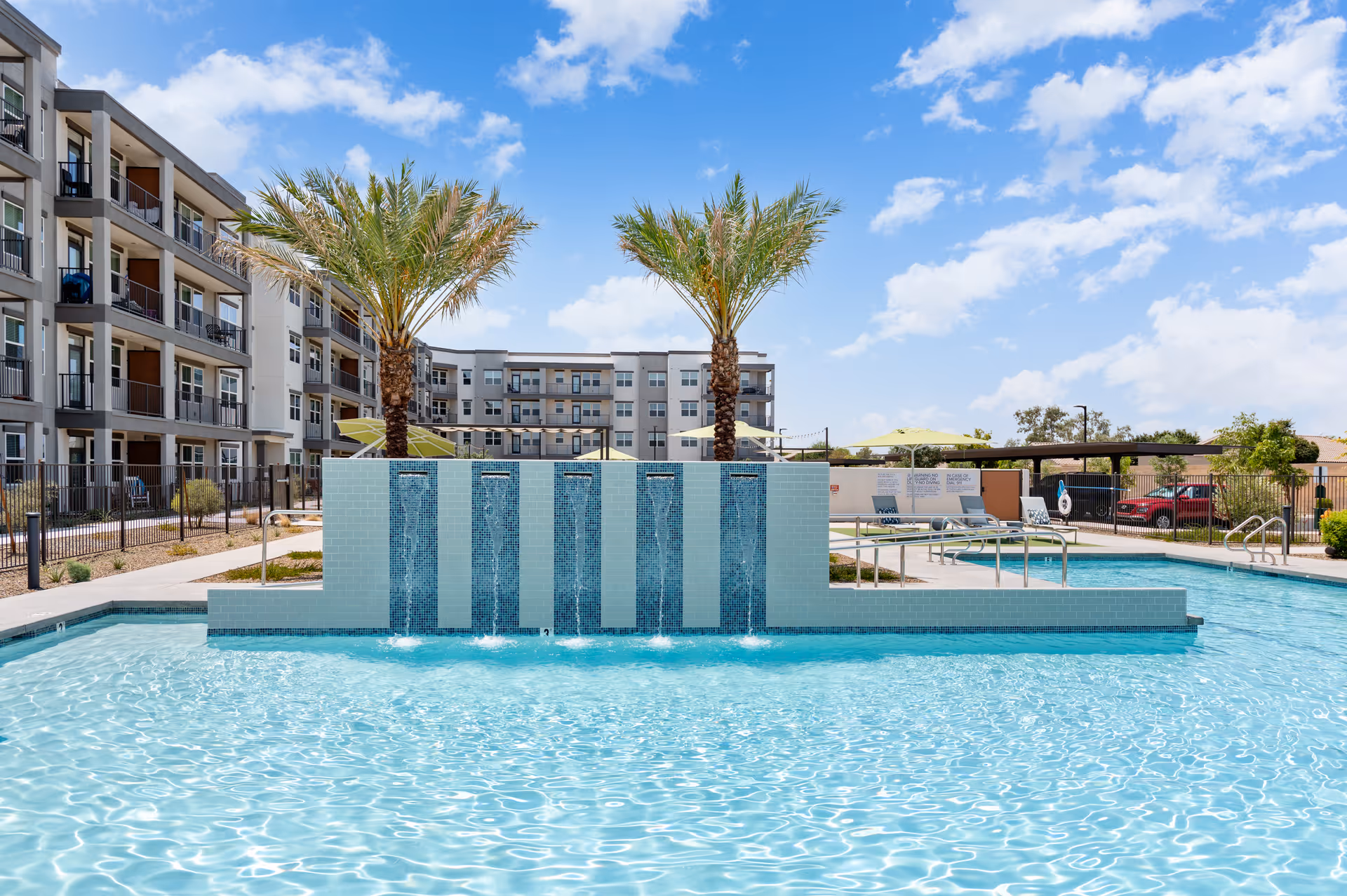Outdoor swimming pool with a tiled water feature, palm trees, and a multi-story residential building in the background.