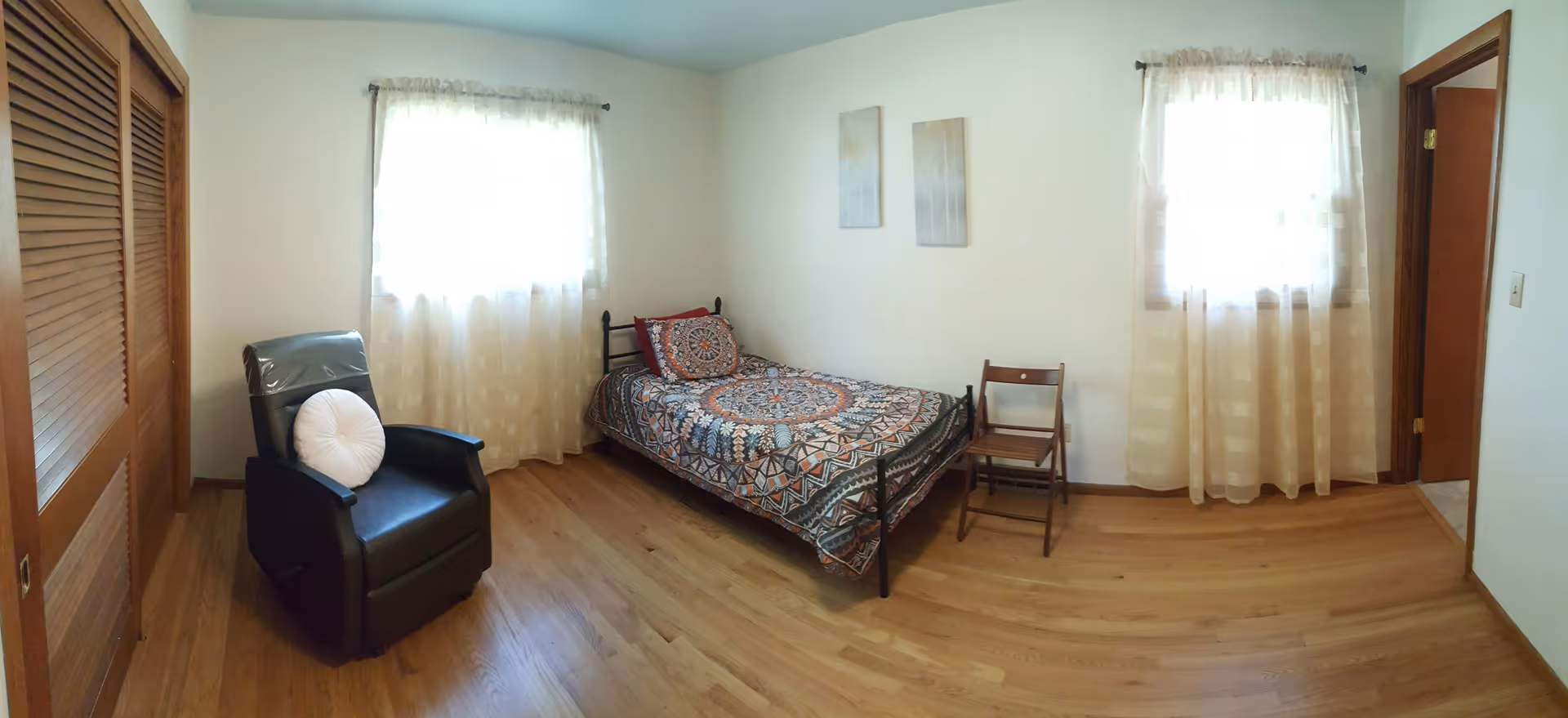 A simple bedroom with wooden flooring, two windows with sheer curtains, a single bed with a patterned bedspread and pillows, a black recliner chair with a round white pillow, a wooden folding chair, and two framed pictures on the wall.