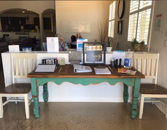 A rustic wooden table with a green base is set against a white half-wall in a bright room with large windows and white shutters. On the table are various items including clipboards, informational pamphlets, a calendar showing May 6, a framed sign that says 'Family,' hand sanitizer, and some small decorative objects. Two white wooden chairs with dark seats flank the table. In the background, a living area with a sofa and people sitting at a table is visible.