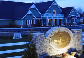 Exterior view of The Courtyards Senior Living facility at dusk, showing a large building with multiple peaked roofs and a well-lit stone sign in the foreground displaying the facility's name.