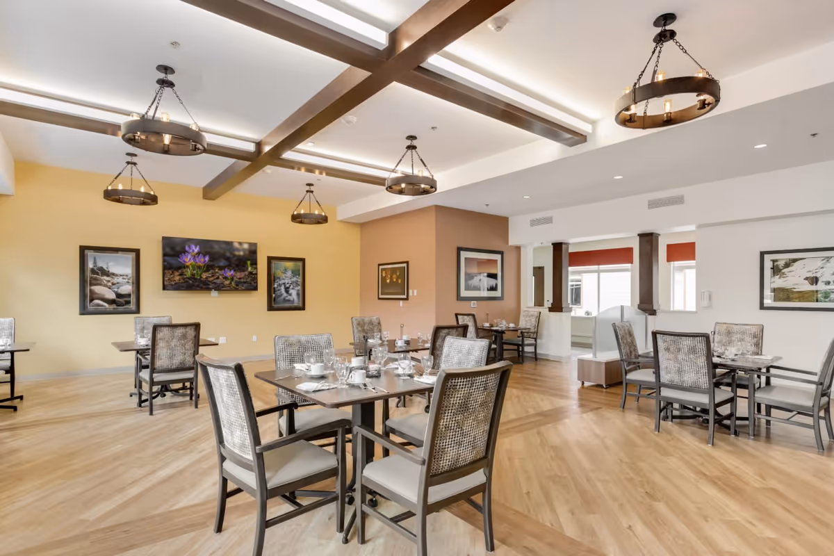 A spacious dining room with several tables and chairs arranged neatly. Each table is set with cups, glasses, and utensils. The room features wooden flooring, light yellow and beige walls adorned with framed nature photographs, and ceiling lights with circular chandeliers hanging from exposed beams.